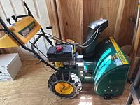 Side view of green and yellow Yard-Man snowblower showing engine, auger housing, wheels with chains and handle