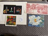 Four art pieces on bed: motel neon sign photo, floral watercolor, abstract watercolor, and French poster.