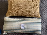 Two pillows stacked, one woven knit pillow in beige on top, striped light blue and white pillow below showing label with lot number.