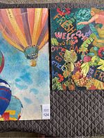 Photo showing two watercolor paintings side by side on a dark quilted surface. The left painting shows multicolored hot air balloons in the sky; the right painting shows a colorful banana garden scene with fruits and text.