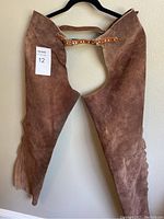 Full front view of brown suede leather chaps hanging on a black hanger.