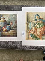 Two canvas paintings laid side by side on a quilted surface, showing full artwork with matting.