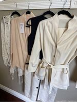 Five vintage clothing items on hangers: cream belted jacket, black and beige sleeveless tops, cream skirt with buttons, and crocheted shawl.