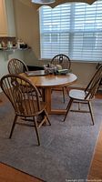 Round wooden dining table with pedestal base and four matching chairs in a room with a window and carpeted floor.
