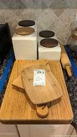 Set of 4 white ceramic canisters with wooden lids, 4 wooden cutting boards, and wooden rolling pin on countertop.