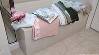 Photo showing various folded linens laid over a bathtub surround, including blankets, towels, and place mats in white, pink, green, and floral colors.