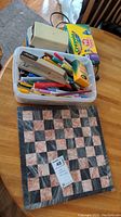 Full view of the plastic container filled with crayons, markers, pencil sharpener, stapler, and the marble chess board underneath on a wooden surface.