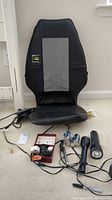 Full view of the black Homedics SBM-200 massage seat cushion with grey panel, power cord, travel adapters, two flashlights, and batteries arranged in front on carpet.