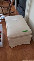 Full view of rectangular ottoman with removable top cushion, showing off-white fabric upholstery and castor wheels.