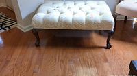 Photo of a rectangular tufted ottoman with ball and claw legs beside a chair on a hardwood floor. Shows top cushioned upholstery with visible stains and wear.