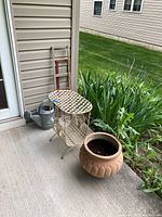Photo showing metal plant stand, ceramic planter filled with dirt, and metal watering can beside green garden plants.