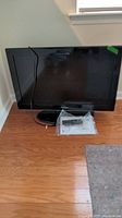 Samsung 32-inch TV on wooden floor with remote and instructions packed on the floor in front of the base.