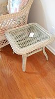 Wicker side table with glass insert top and woven lower shelf placed near wicker armchair on wooden floor