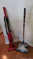 Two vacuums side by side against wall on wood floor: red upright Dirt Devil with cord and silver/blue cordless Shark sweeper.