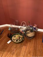 Wide view showing assortment of wicker baskets, glass vases, ceramic platters with faux fruit and small decor items arranged on hardwood floor.