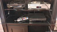 Image showing four electronic devices arranged in a shelf unit, including a silver and black Memorex VHS/DVD player, black Sony VCR, black RCA VCR, and a black power center with cables attached.