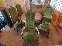 Round wooden dining table with six emerald green-upholstered chairs surrounding it, showing details of wood grain and carved legs.