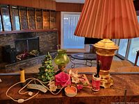 Wide view of all items on brown table surface with fireplace and seating area in background, showing lamp, ceramic tree, green glass lamp, floral decor, lint roller, remote control, and ashtray.
