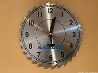Mastercraft wall clock with circular saw blade design, black tool-shaped hour and minute hands, red second hand, black numerals, and blue Mastercraft logo on silver metal face.