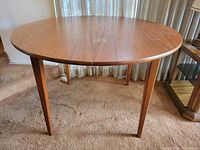 Angled view of the circular teak dining table showing the tabletop and legs on carpeted floor.
