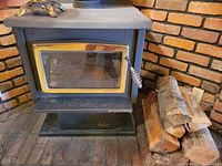Front view of Osburn wood stove showing glass door with metal spring handle and brass/gold trim. Stove is set on brick surface with decorative metal element on top and firewood beside it.