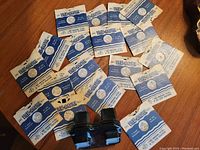 Photo showing multiple View-Master slides in blue and white paper sleeves spread out on a wooden surface with the black stereoscopic viewer present.