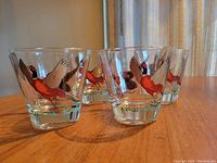 Four clear glasses arranged on wood table, each glass with a multi-colored pheasant in flight illustration.