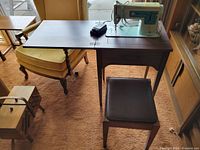 Singer Model 347 sewing machine mounted in wood cabinet with fold-out extension table and matching wooden stool, shown together in a room with carpet flooring and armchair nearby.