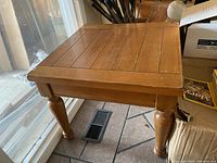 Side angle of a square wooden side table showing panel top and turned legs, placed on a tiled floor near a window.
