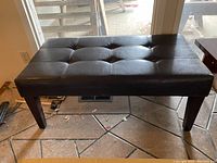 Full view of rectangular black leather bench with tufted seat and wooden legs.