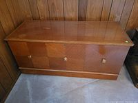 Full view of the closed wooden blanket chest against a paneled wall, showing the midsize rectangular shape and wood veneer finish.