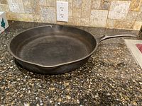Full side view of the round Wagner cast iron pan with handle resting on a granite countertop, showing the pouring spouts on opposing sides and solid handle.