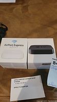 Photo showing both Apple AirPort Express Base Station and Apple TV adapter boxes placed side by side on a brown surface, along with a paper listing the items.