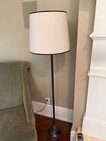 Full view of the floor lamp next to a chair and wall outlet, showing the tall black metal stand and white drum lampshade with black edge.