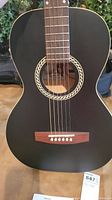 Front body of black Art & Lutherie Ami Wild Cherry acoustic guitar showing cream binding, rope rosette, rosewood bridge and six steel strings.