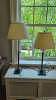 Two adjustable height lamps placed on a white cabinet in front of a window with green trees outside. One lamp is set lower, the other taller.