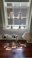 Five chrome lamps arranged by a window and on the floor; two with beige shades on windowsill, two chrome desk lamps on floor with dome shades, one taller lamp with cream shade on floor.