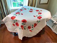 Full view of white quilt with red floral wreath design and scalloped edges placed on table under window.