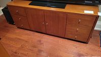 Front view of the wood television console showing six drawers and two doors with silver handles, sitting on a hardwood floor with a television on top.