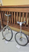 Two unicycles side by side, one slightly taller with larger front wheel and one shorter with smaller wheel, both chrome framed with black seats and pedals.
