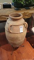 Full view of large pottery urn with multiple handles on black metal stand on hardwood floor near furniture.