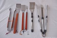 Two sets of BBQ utensils laid out on a white surface, showing metal-handled and wood-handled tools including tongs, forks, and spatulas.