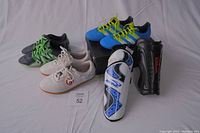 Four pairs of boys sports shoes with two pairs of shin guards laid out on floor, showing colors, sizes and brands.