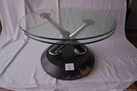Front-side angle view showing the clear round glass top, three metal crossbar supports, and the black circular base with visible wear.