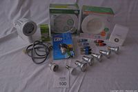Assorted LED light fixtures and bulbs laid out on a flat surface, including floodlight, ceiling fixtures in packaging, spotlight bulbs, motion sensor light, and small box of bulbs.
