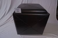 Closed cube-shaped black leather or leather-like storage ottoman showing diagonal stitch details on sides.