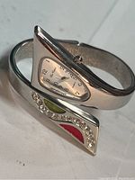 Close-up shot of the bracelet watch showing the decorative side with rhinestones and colored enamel accents in red and green, alongside the metallic body.