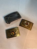 Three belt buckles shown together: two gold-tone metal Gypsy Tour Award buckles and one dark gray leather buckle