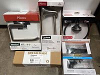 Photo showing the boxed Pfister Venturi Toilet Paper Holder, Speakman door knob box, and three boxed towel rings by Delta and Defiant.