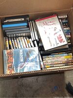 Box containing over 80 used DVDs in cases, visible titles include 'Brick' and 'Meet the Parents'. Multiple rows of DVD cases stacked inside a cardboard box.